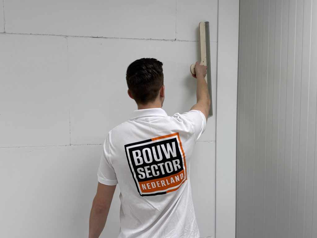 Van dichtbij: stukadoor met Bouwsector-logo op zijn shirt zet een brede spaan tegen de muur