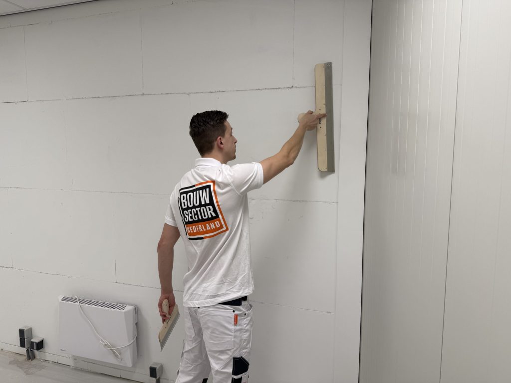 Stukadoor in wit werkpak werkt met spaan en plakspaan aan een wand boven een elektrische kachel