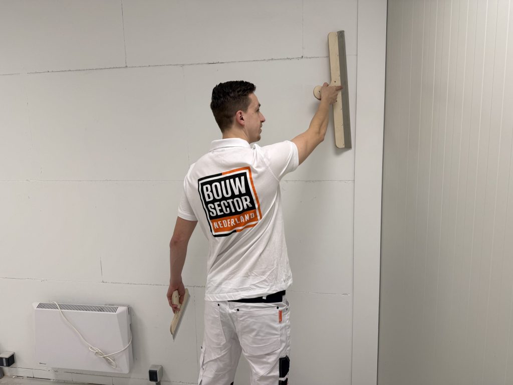 Stukadoor van Bouwsector Nederland strijkt een muur glad met een brede spaan en houdt een plakspaan in zijn andere hand